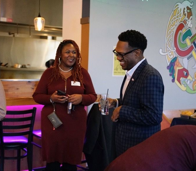 Two people in professional attire smiling and talking in a restaurant with a mural on the wall.