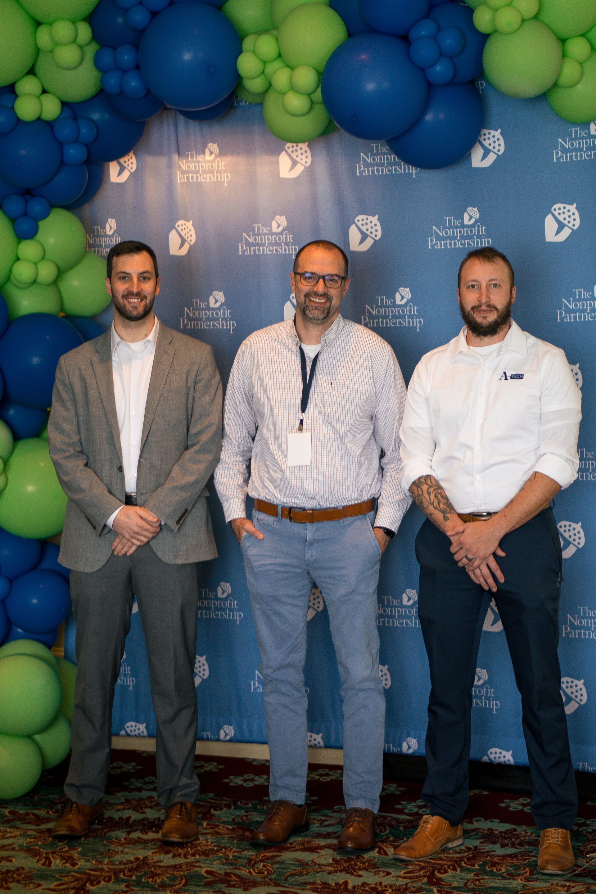 Three men stand smiling in front of a blue banner with a green and blue balloon arch at an indoor event.