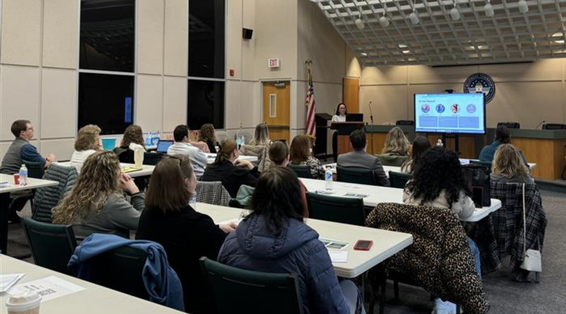 Attendees sit at tables in a room, facing a screen showing a presentation while a speaker stands at the front.