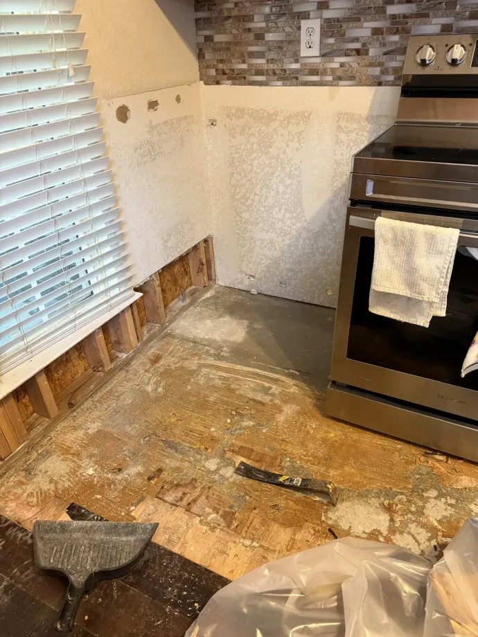 Kitchen floor under renovation with exposed subfloor, a stove, and a dustpan near the window blinds