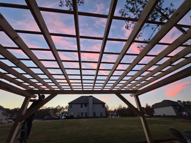 Wooden pergola frame at sunset overlooking a house and open yard under a pink sky