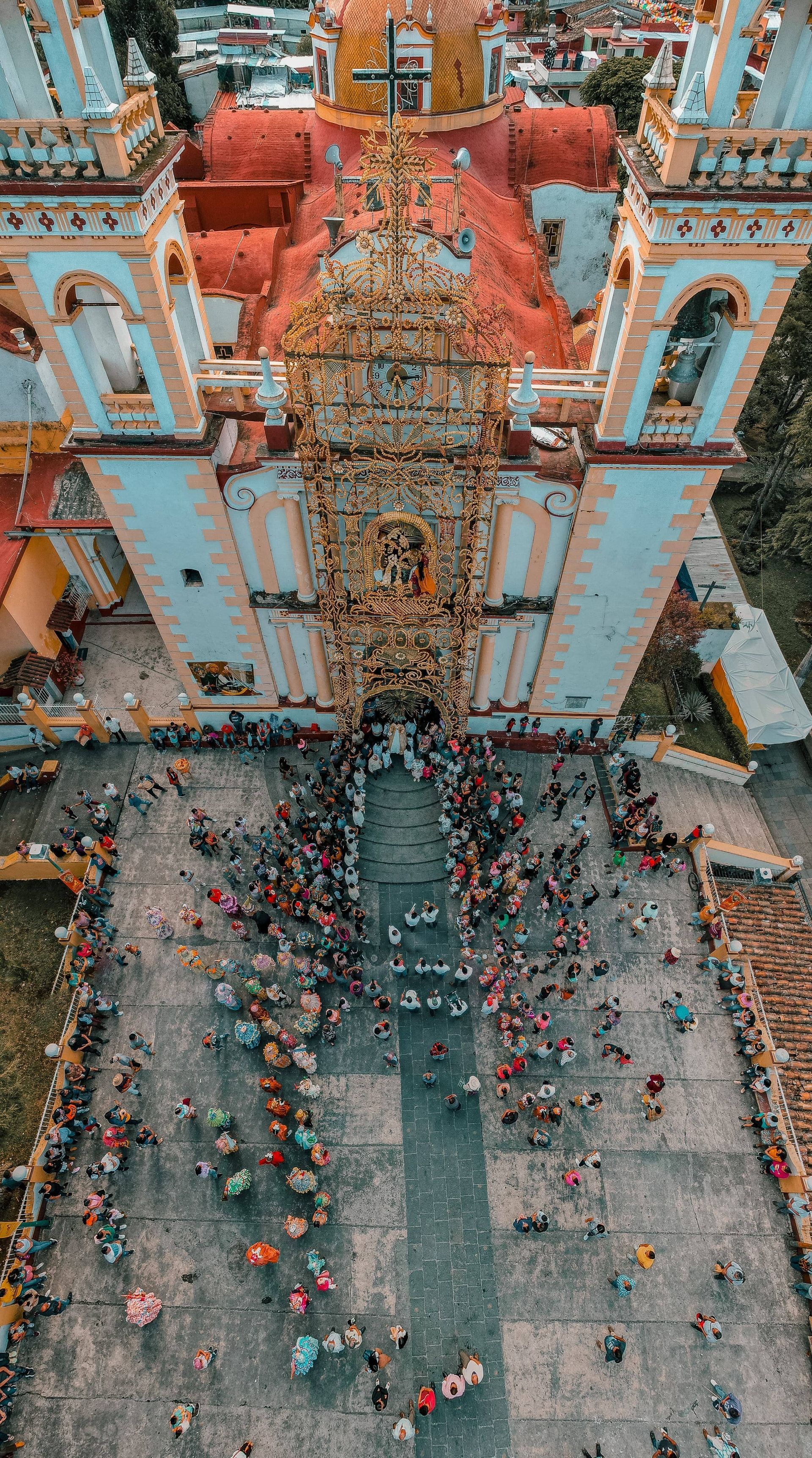 Crowded street procession outside a colorful church, with a tall decorated tower and gathered people below.