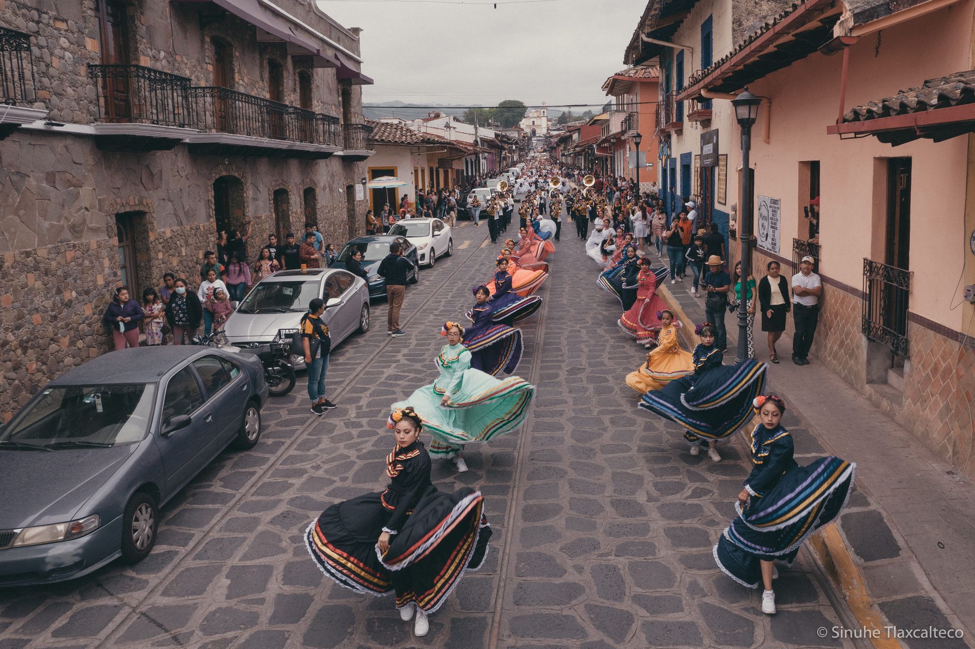 Dancers in colorful skirts perform a street parade along a cobblestone road lined with spectators and buildings