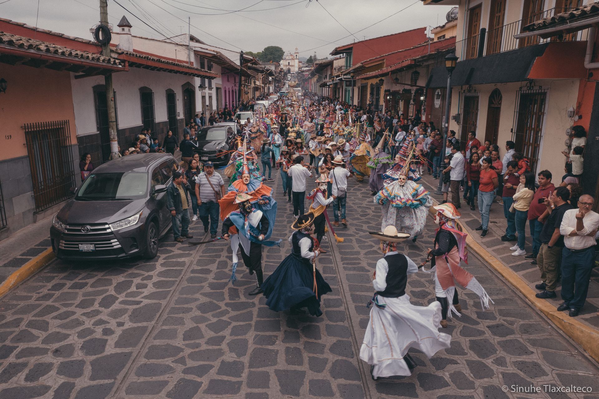 Desfile callejero con gente vestida con coloridos disfraces en una calle empedrada bordeada de edificios.