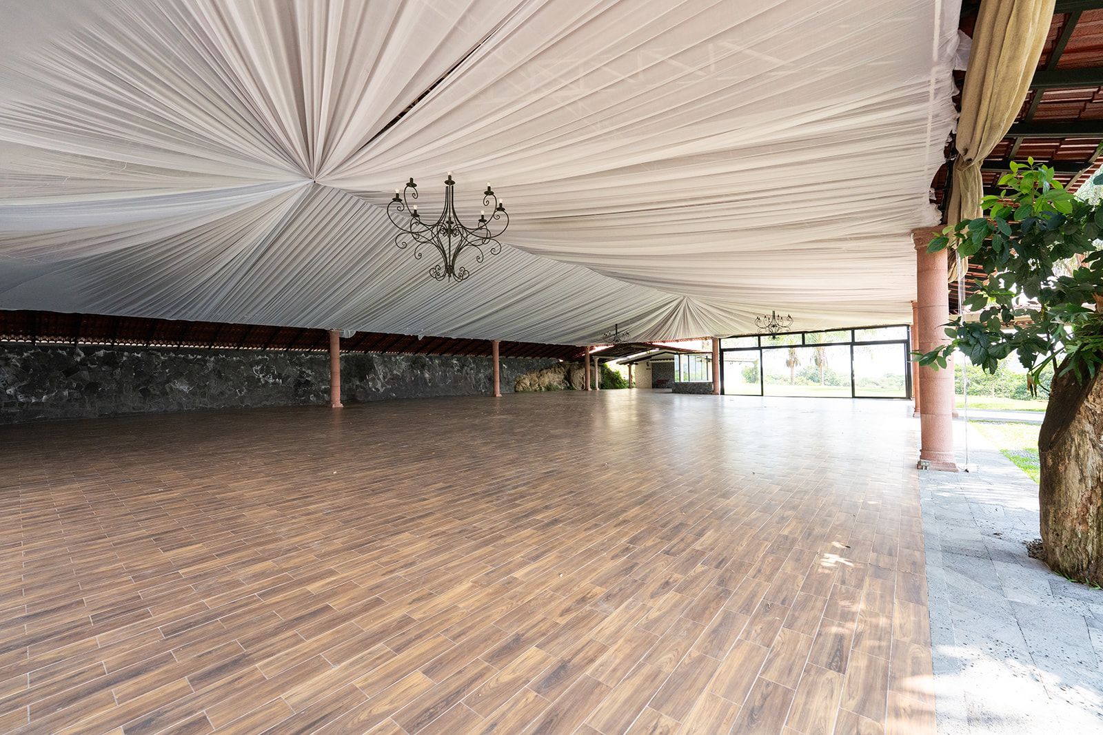 Spacious outdoor pavilion with a wooden floor, draped white ceiling, and open sides overlooking greenery