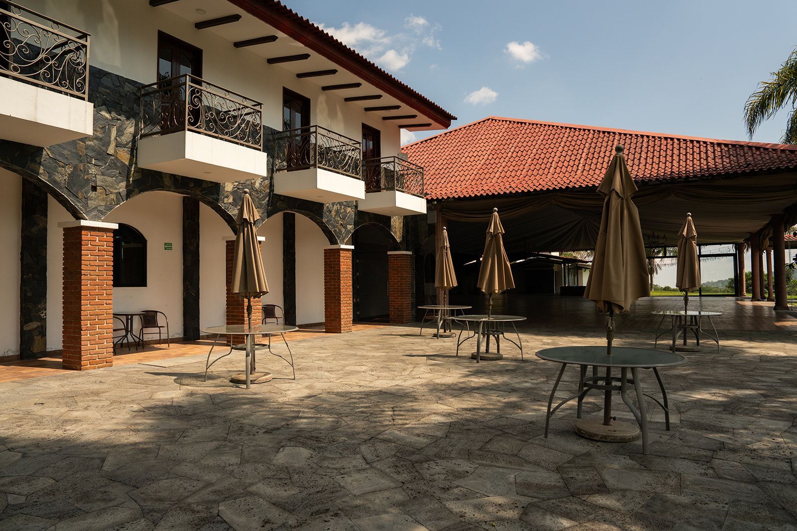 Courtyard with stone paving, outdoor tables, umbrellas, and a two-story building with balconies and arches