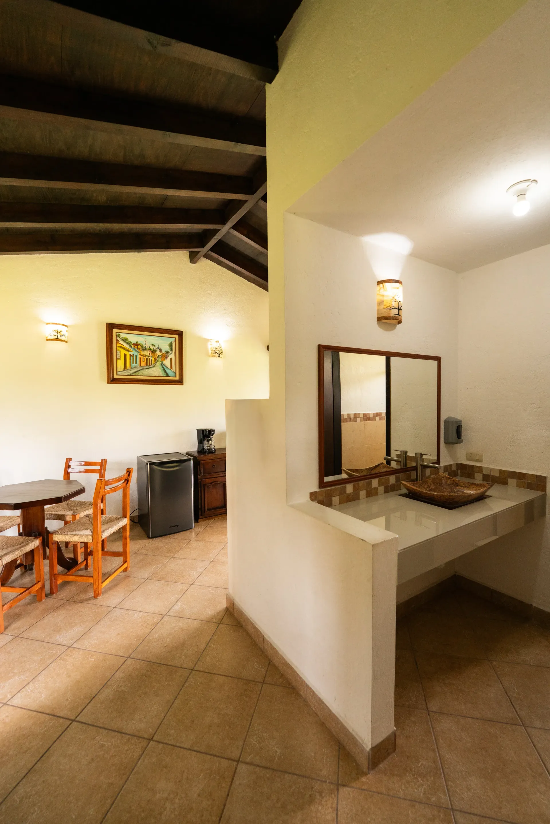 Comedor luminoso con suelo de baldosas, mesa y sillas de madera, cuadros en las paredes y tocador con espejo y lavabo.