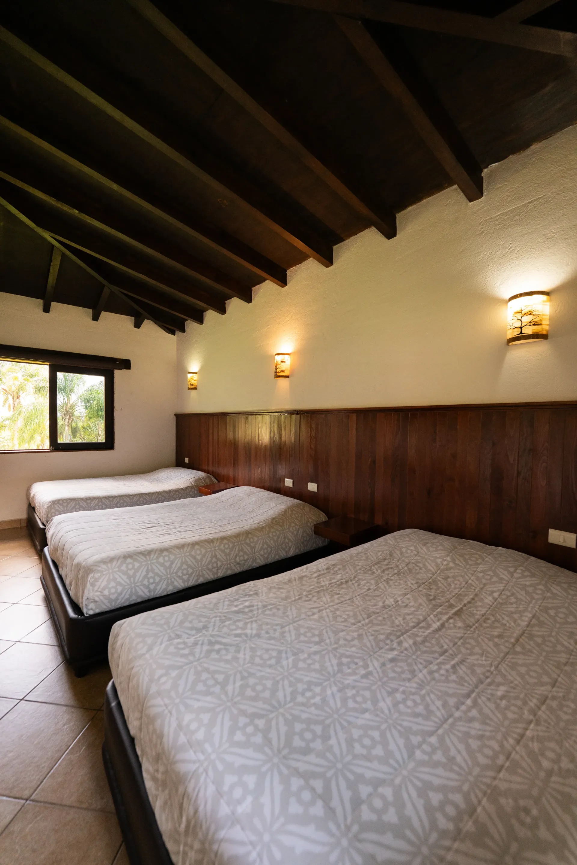 Dormitorio rústico con tres camas bien hechas, paredes revestidas de madera, techo inclinado y apliques de pared.