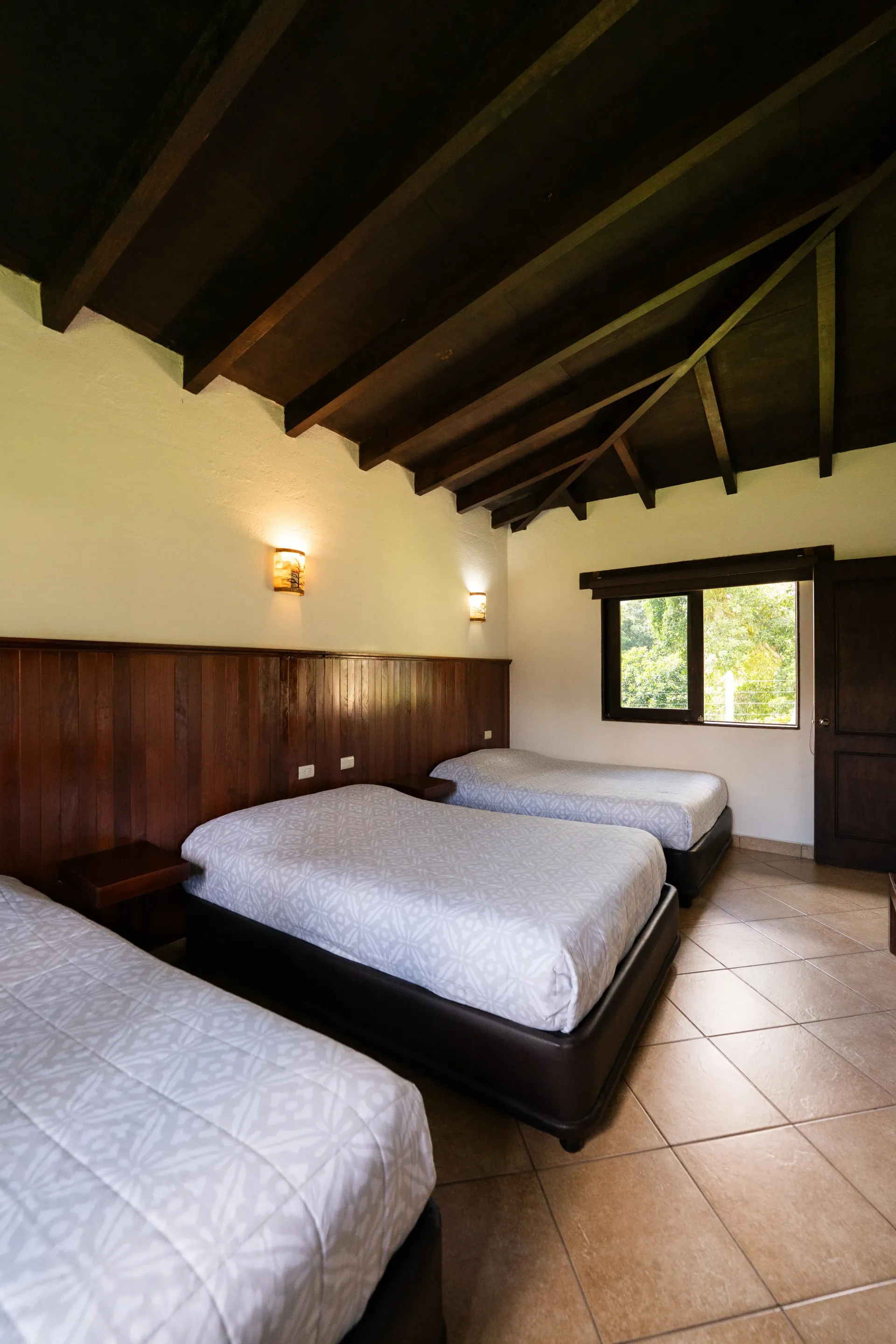 Pequeña habitación rústica con dos camas, paredes revestidas de madera y suelo de baldosas cerca de una ventana y una puerta.