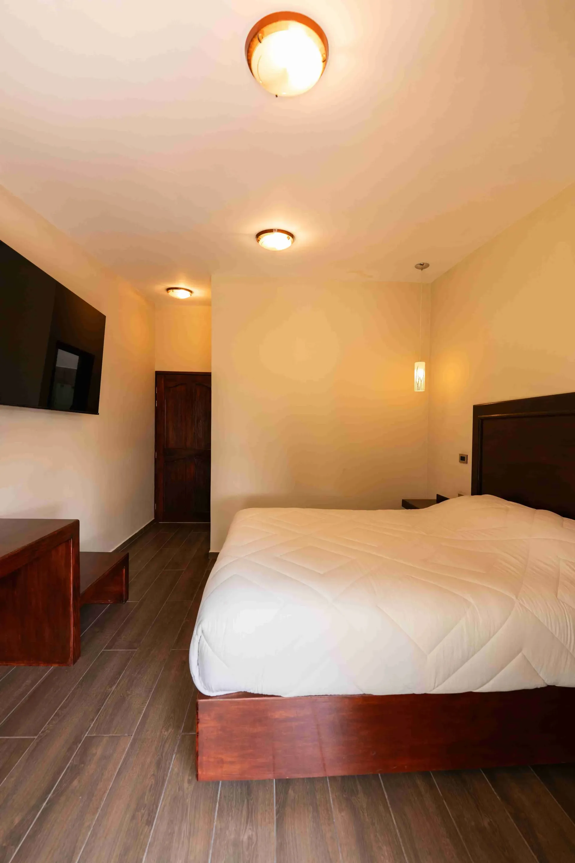 Una pequeña habitación de hotel de color beige con cama de madera, televisor, cómoda y luces de techo.