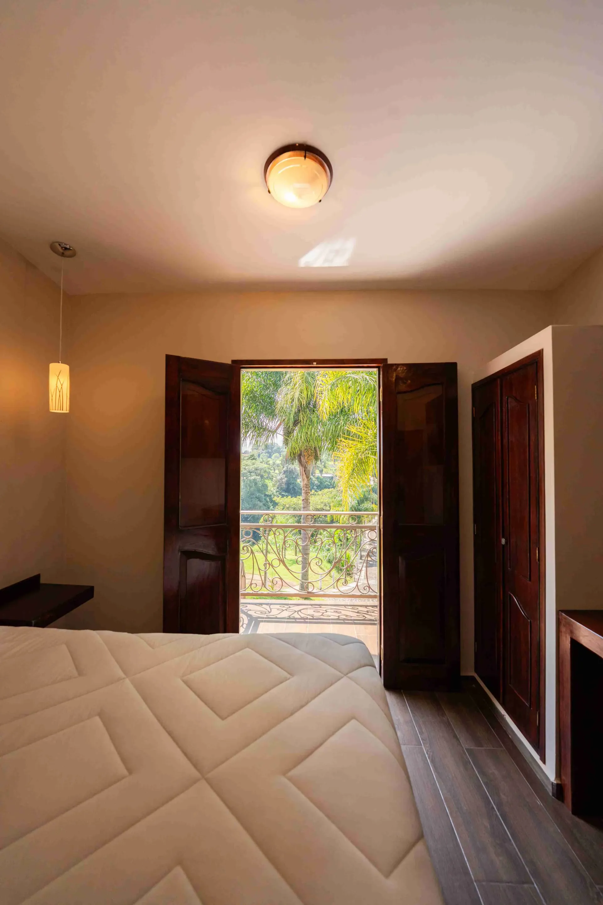 Dormitorio con puertas abiertas que dan a un balcón, una cama blanca en primer plano y luz solar que entra a raudales.