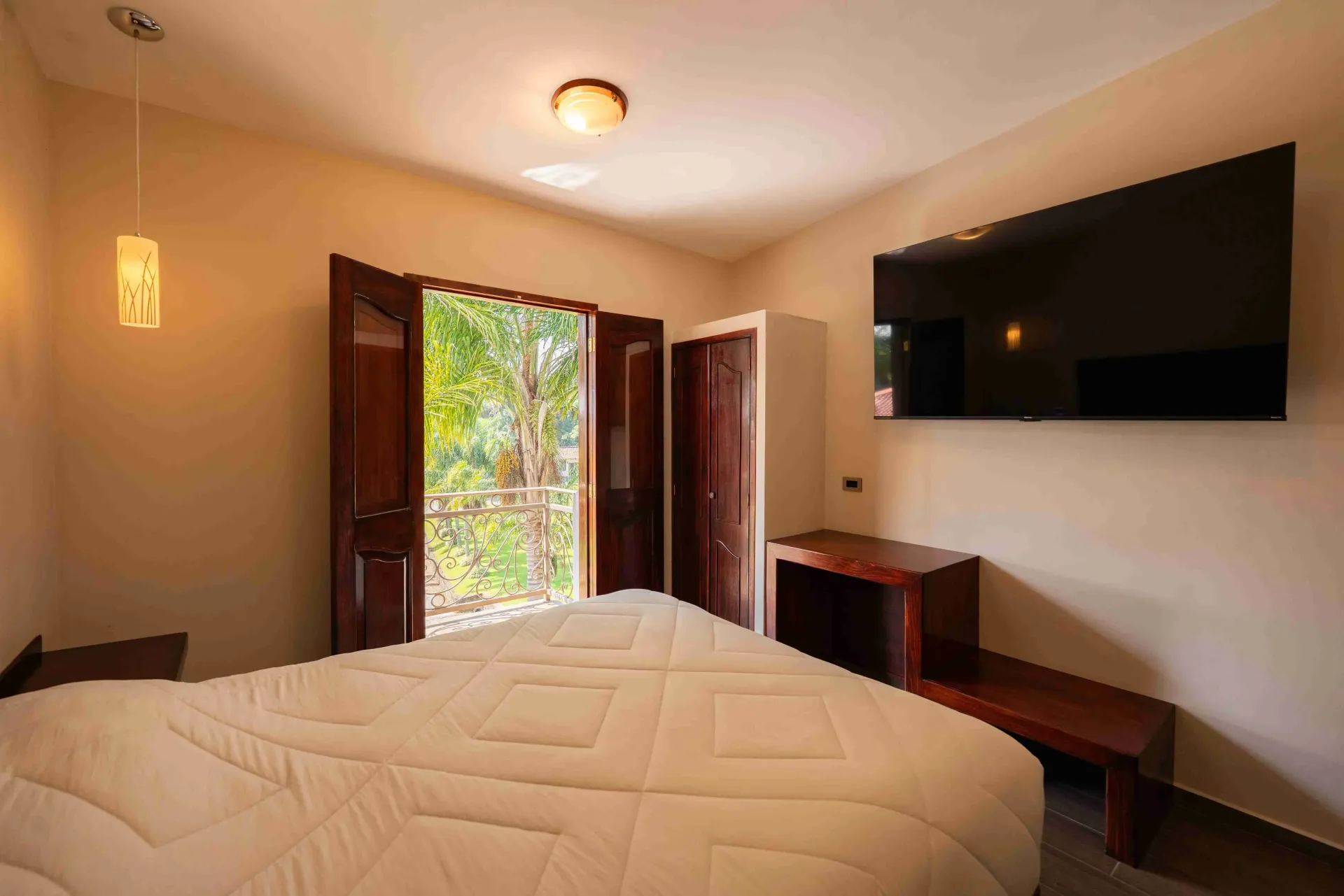 Habitación con cama sin hacer, puertas de balcón abiertas y televisor montado en la pared.