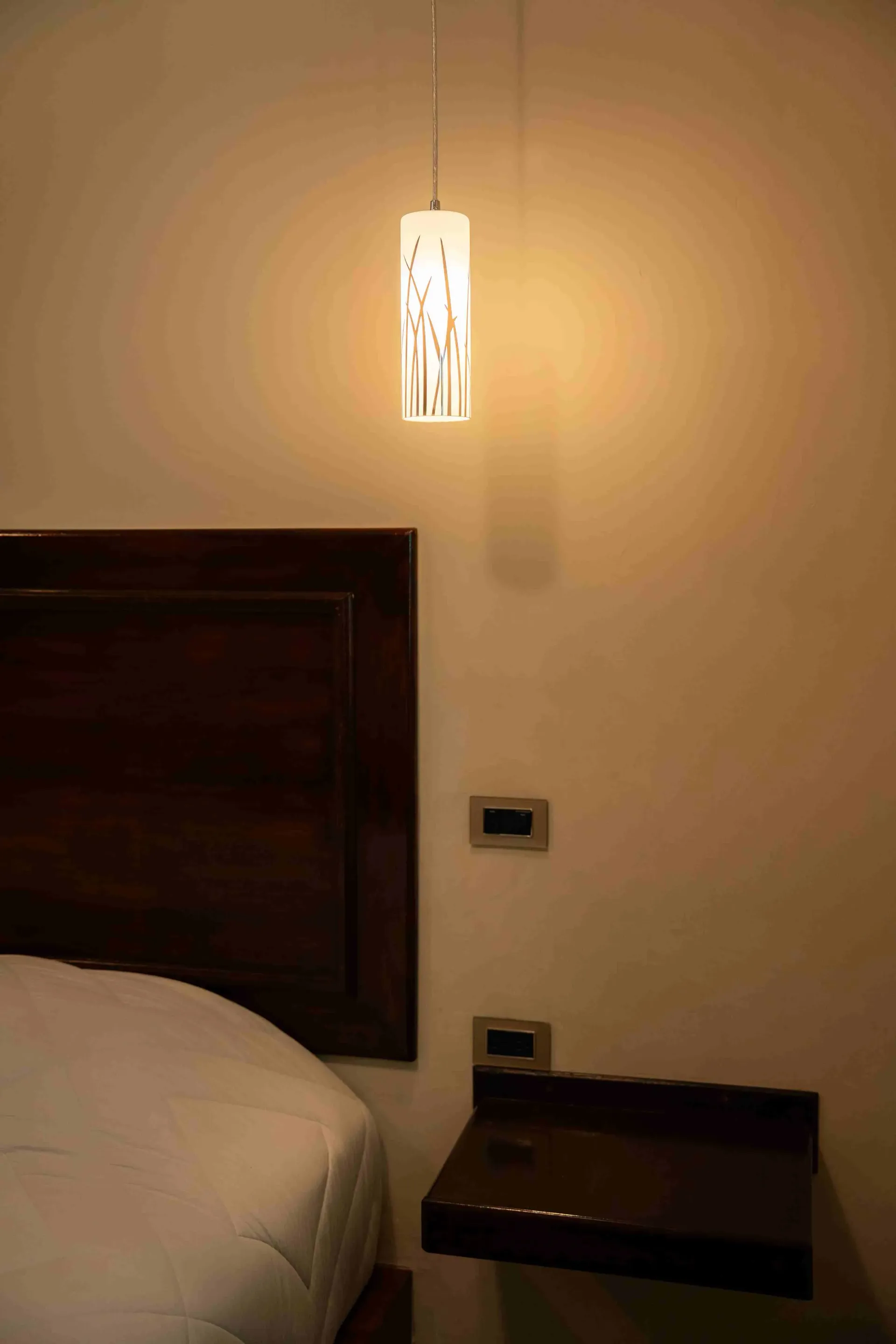 A softly lit bedroom corner with a bed, wall lamp, and dark bedside table.