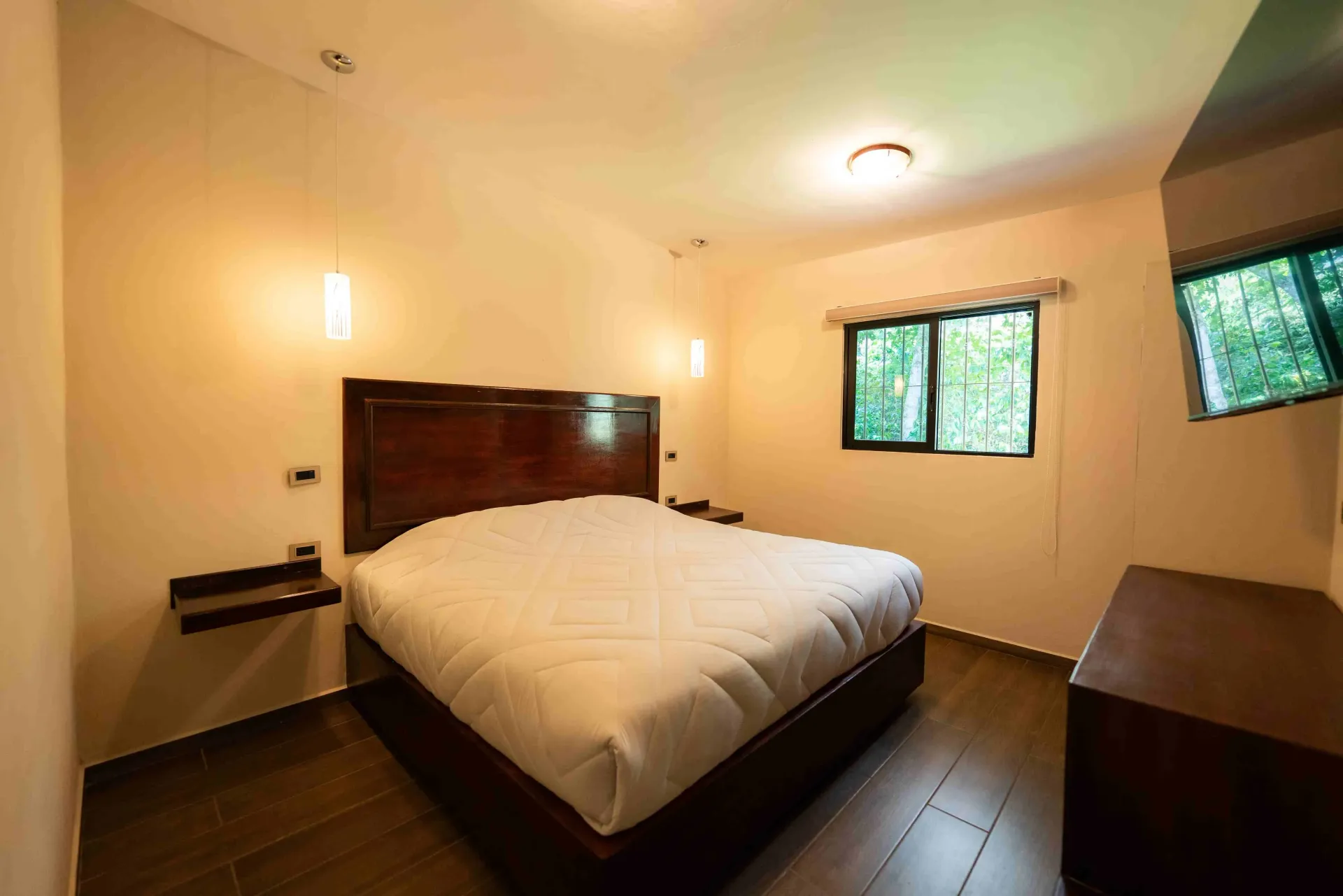 Habitación moderna con una cama grande, iluminación cálida, suelo de madera oscura y dos ventanas con vistas verdes.