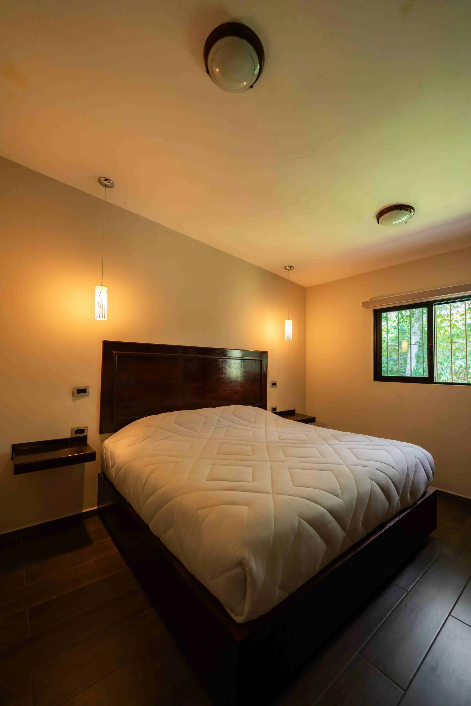Bedroom with a large bed, dark wood headboard, warm wall lights, and a window showing greenery.