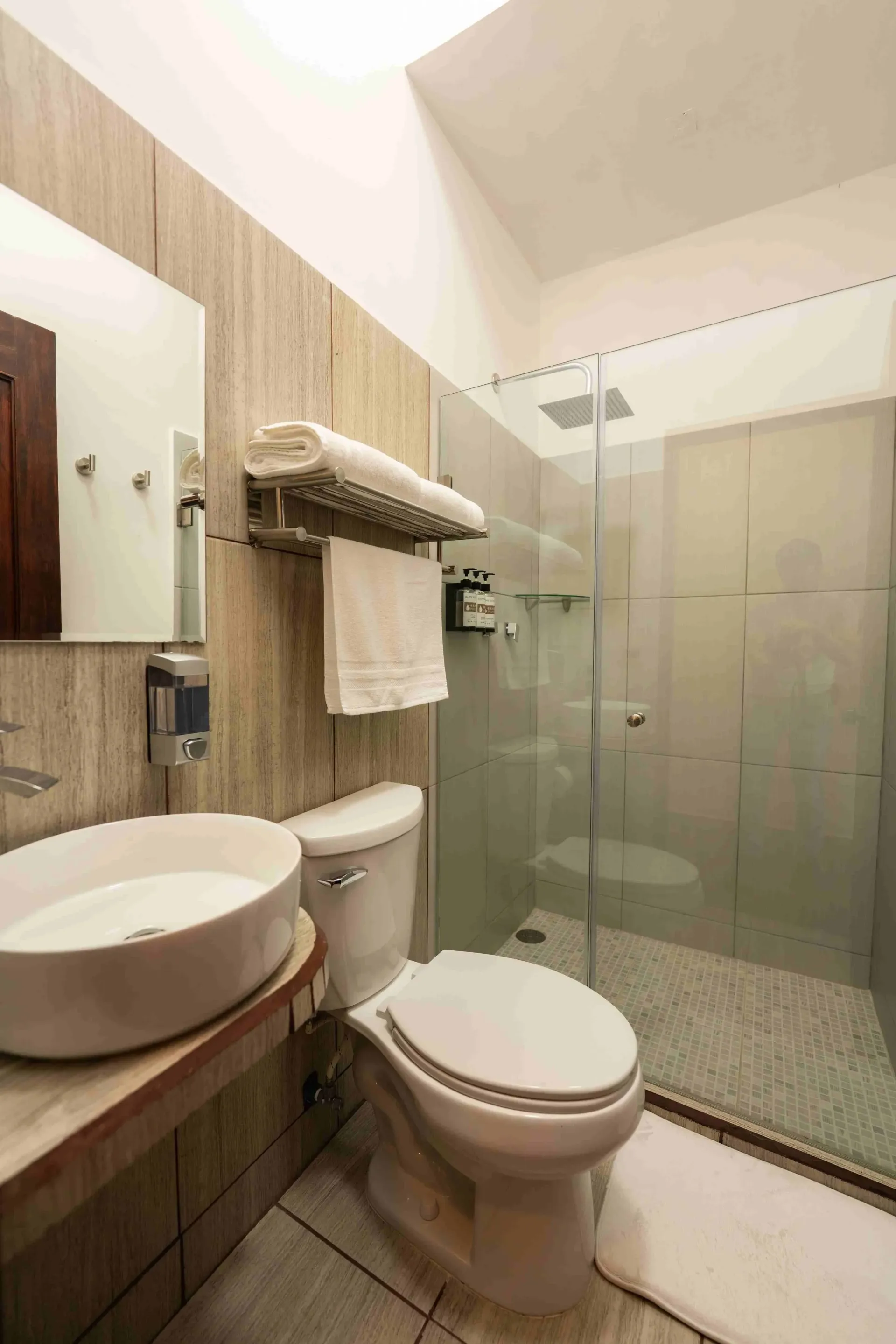 Baño compacto de color beige con lavabo, inodoro y ducha a ras de suelo con mampara de cristal.