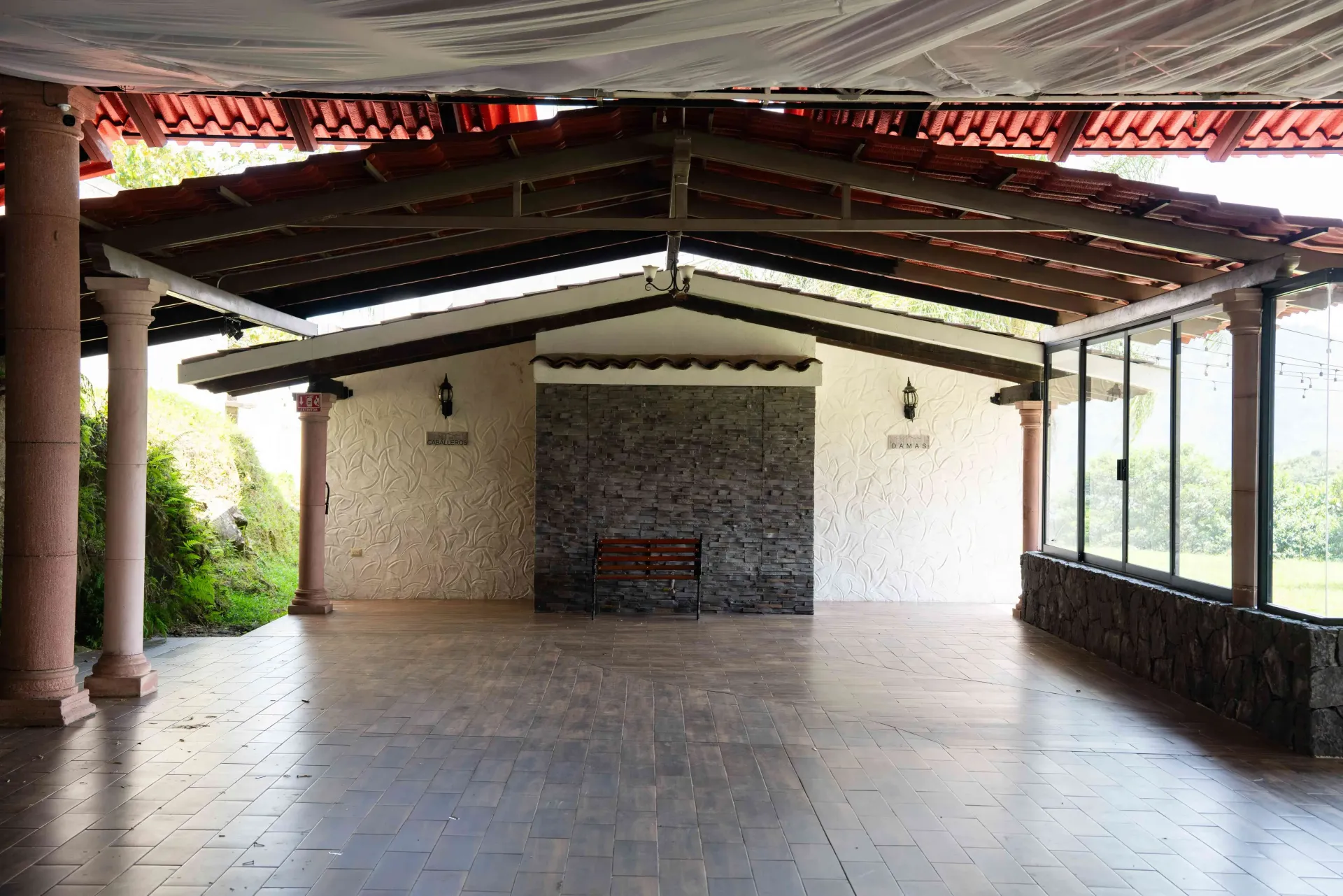 Covered outdoor patio with tiled floor, stone wall, and open sides overlooking greenery