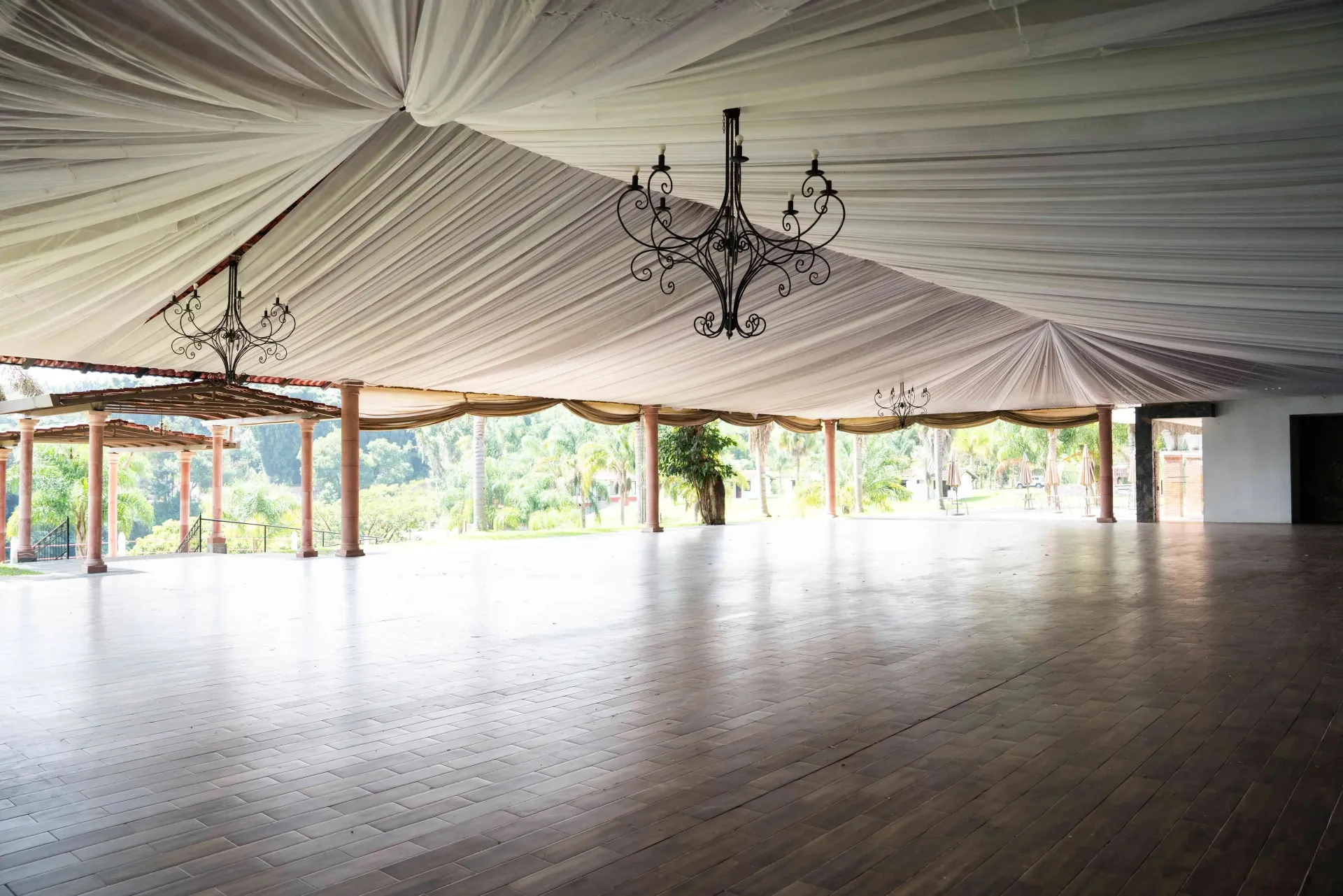 Empty indoor pavilion with polished floor, draped ceiling, and open sides overlooking greenery