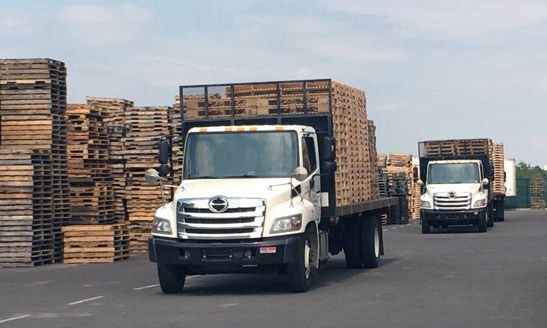 Two white trucks hauling stacked wooden pallets in an industrial yard