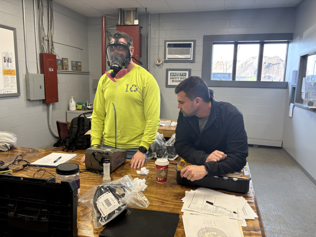 Two people in a workshop stand by a table with tools, papers, and drinks; one wears a neon yellow top.