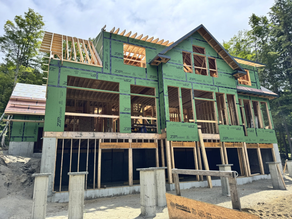 Green house under construction with exposed framing, roof trusses, and concrete foundation in a wooded area