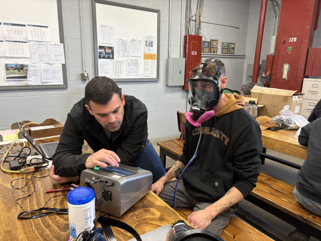 Two people in a workshop, one adjusting equipment while the other wears a respirator mask and pink gloves.