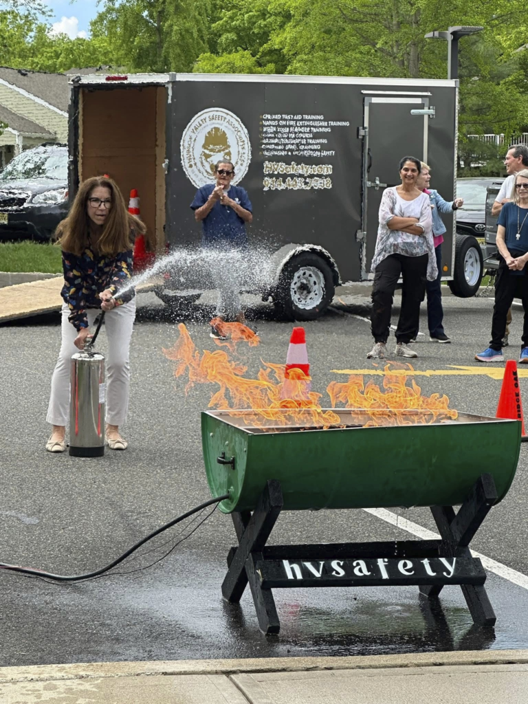 Outdoor fire safety demo with a woman using a fire extinguisher on flames in a metal bin