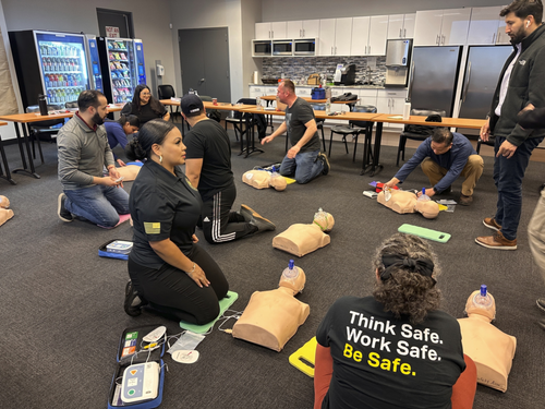 People kneel in a classroom practicing CPR with manikins and training kits on the floor.