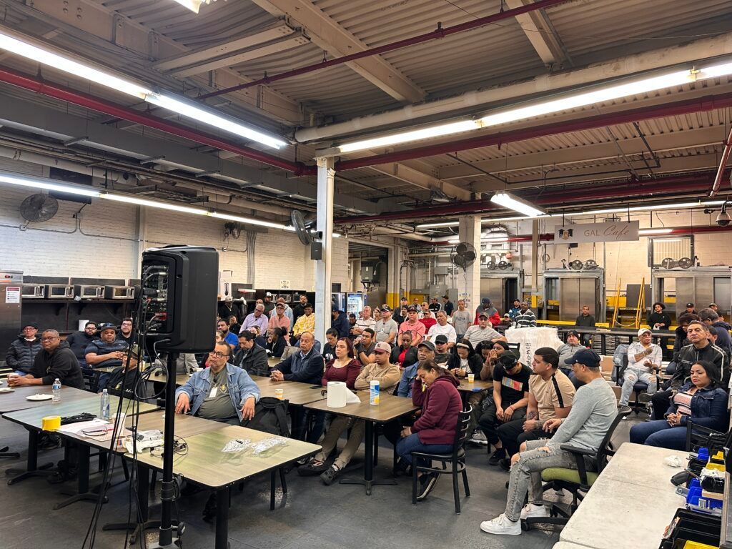 People seated at long tables in a busy industrial workshop, watching a presentation.
