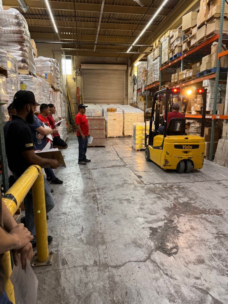 Warehouse aisle with a yellow forklift and several workers standing along the left side.