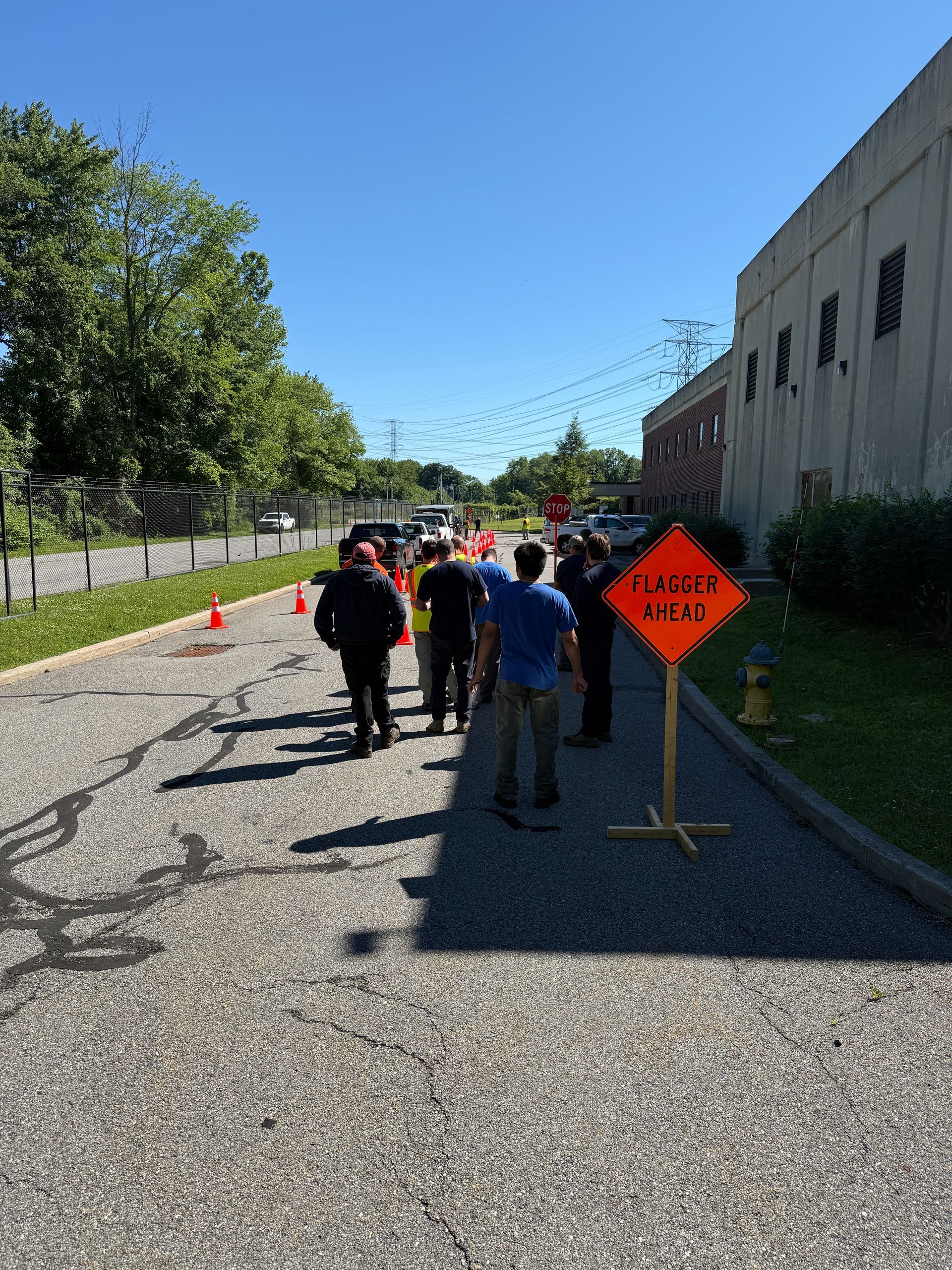 Workers and cones line a sunny sidewalk beside parked trucks and a building.