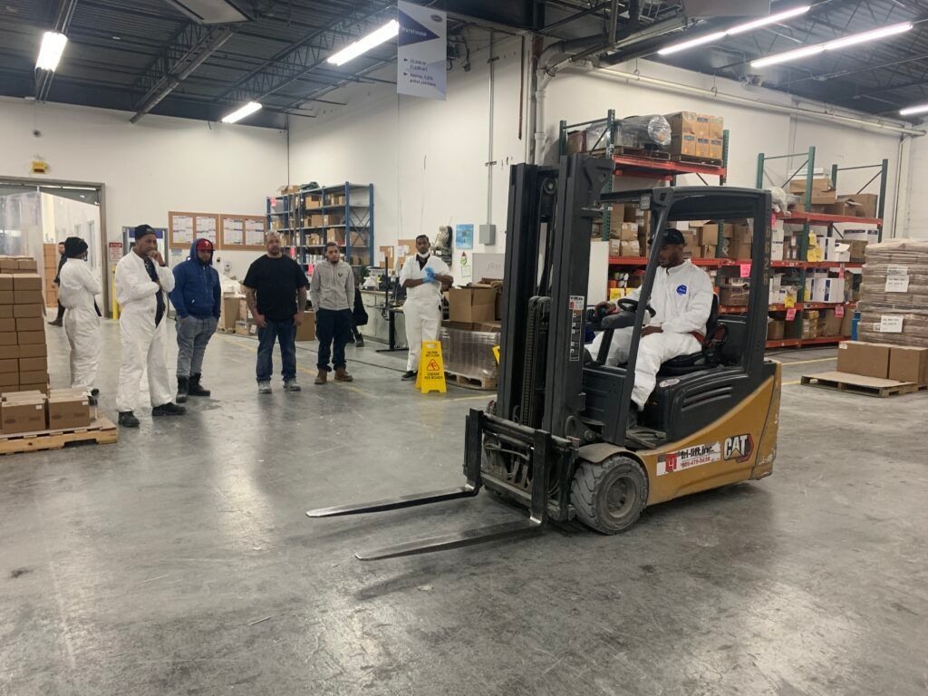 Workers and a forklift in a warehouse aisle with shelves and safety gear nearby