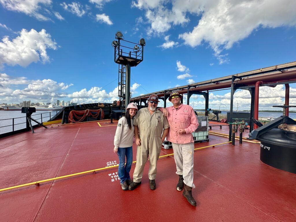 Three people standing on a red ship deck under a blue sky with clouds.