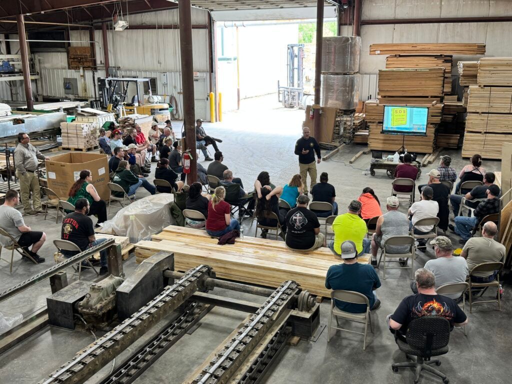 People seated in a workshop listening to a presentation near stacks of lumber and a large screen.