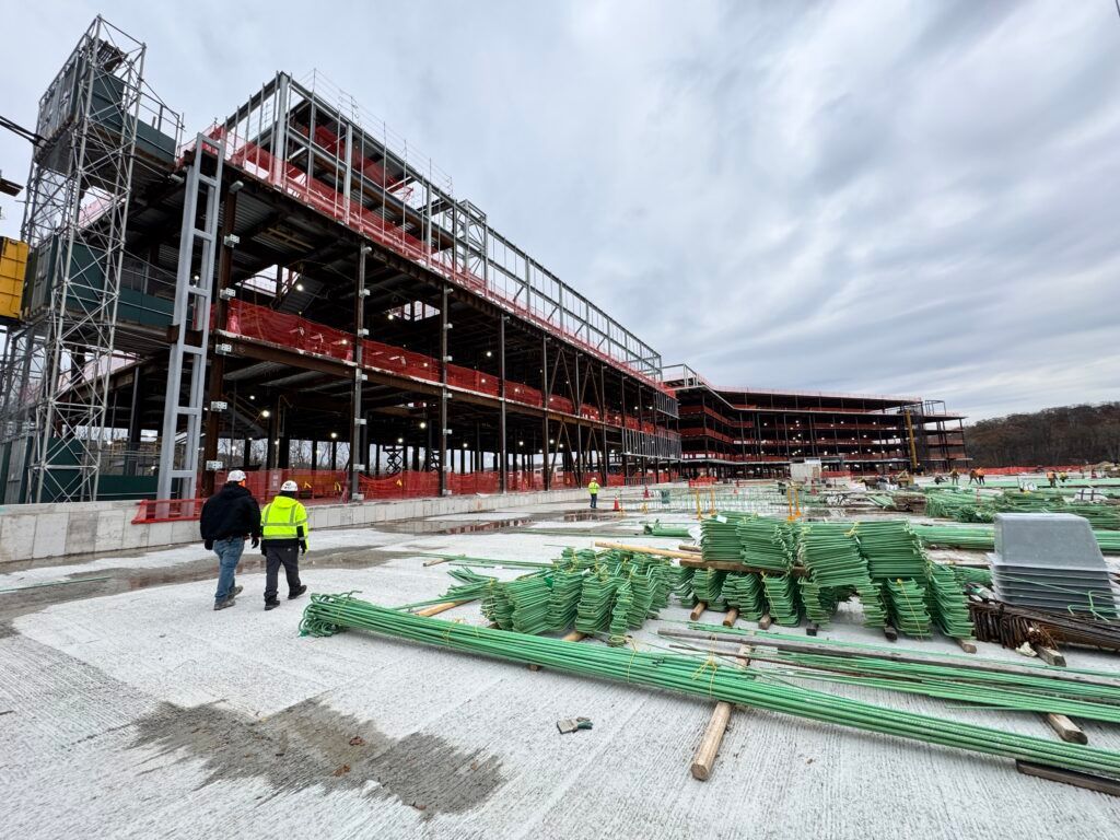 Construction site with steel framework, workers, and stacks of green materials under a cloudy sky