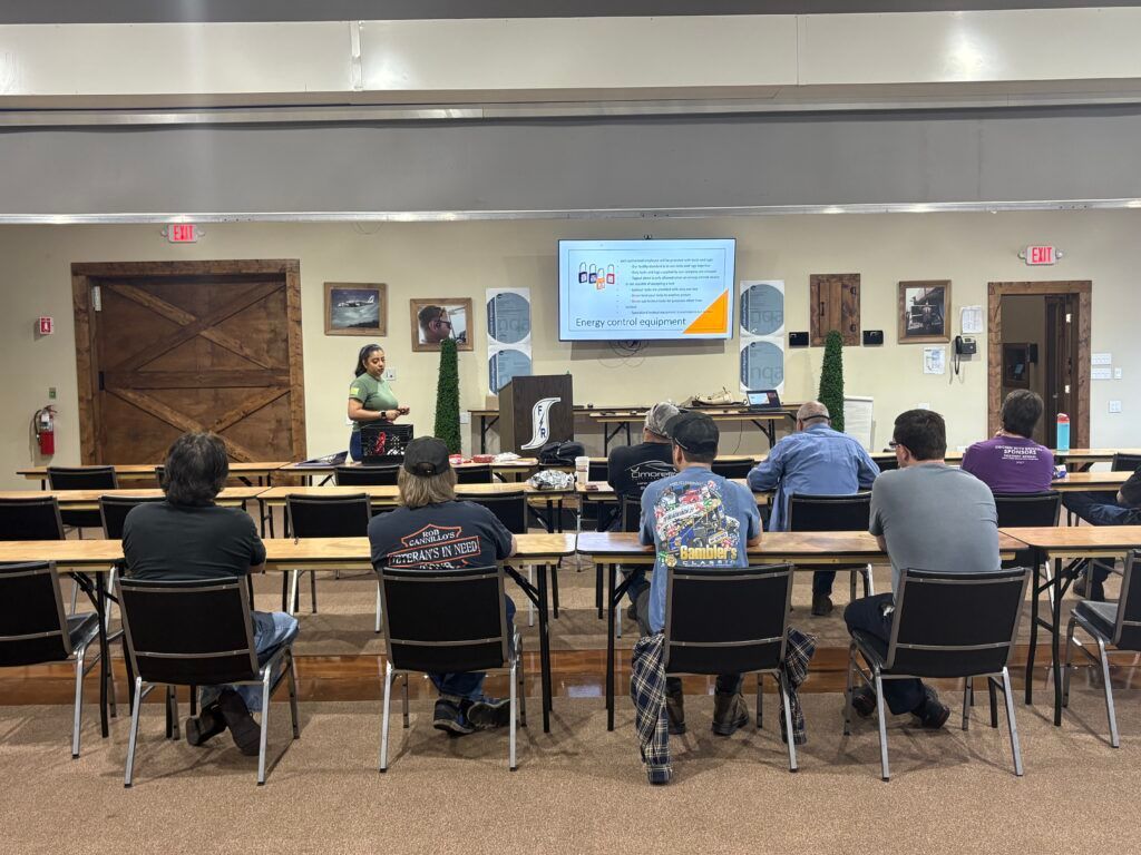 People seated in a conference room watching a speaker at the front near a screen and piano.