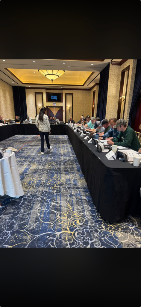 Conference room with attendees seated at long tables while a presenter stands at the front.