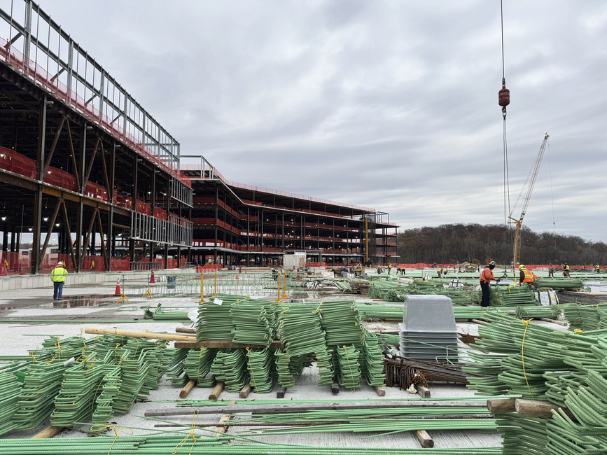 Construction site with steel frame building, crane, and workers on a concrete slab with green materials stacked in foreground