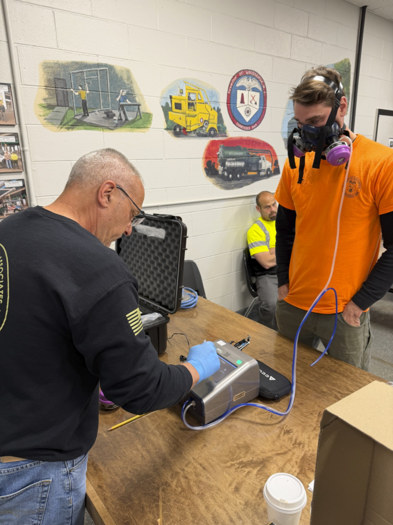 Two people in a workshop testing a device on a wooden table, with plaques on the wall behind them