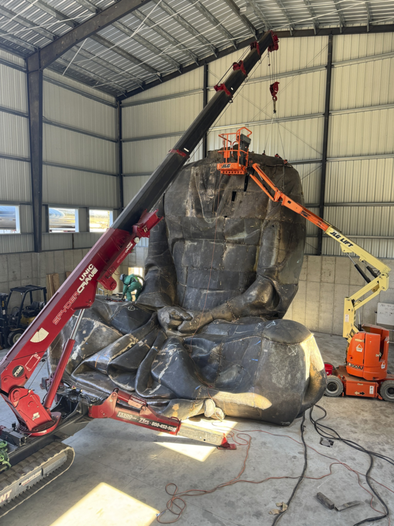 Large black wrapped sculpture being rigged in a warehouse by a crane and workers