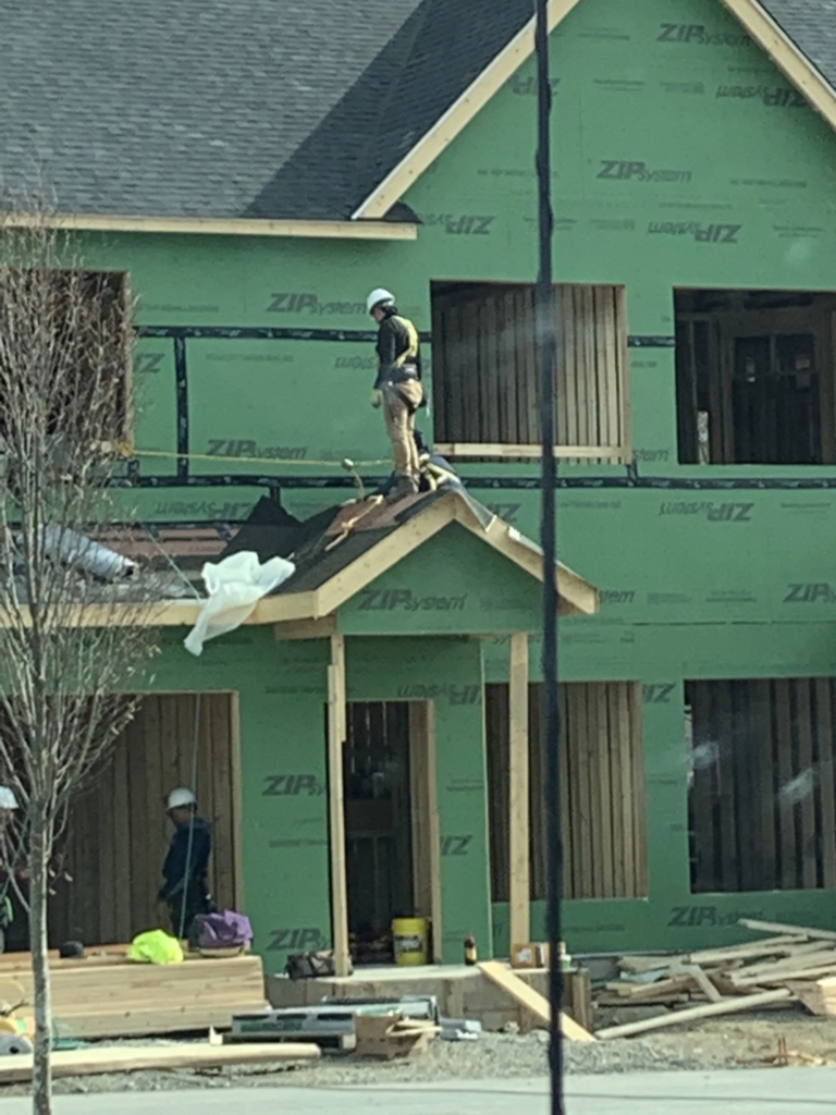 Construction workers on a house roof with green wrap and lumber in front