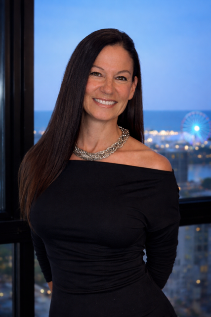 Woman in a black off-shoulder dress smiling by a window with a city skyline at dusk