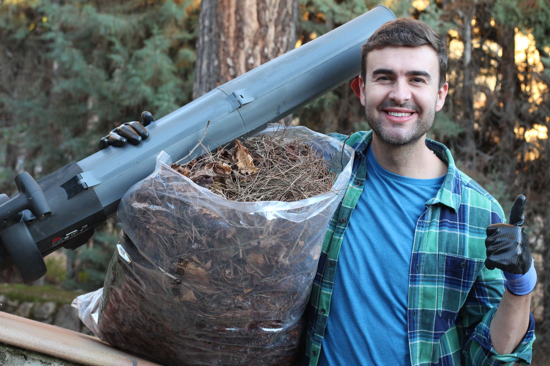 Man holding a leaf blower and large bag of leaves outdoors, smiling and wearing work gloves