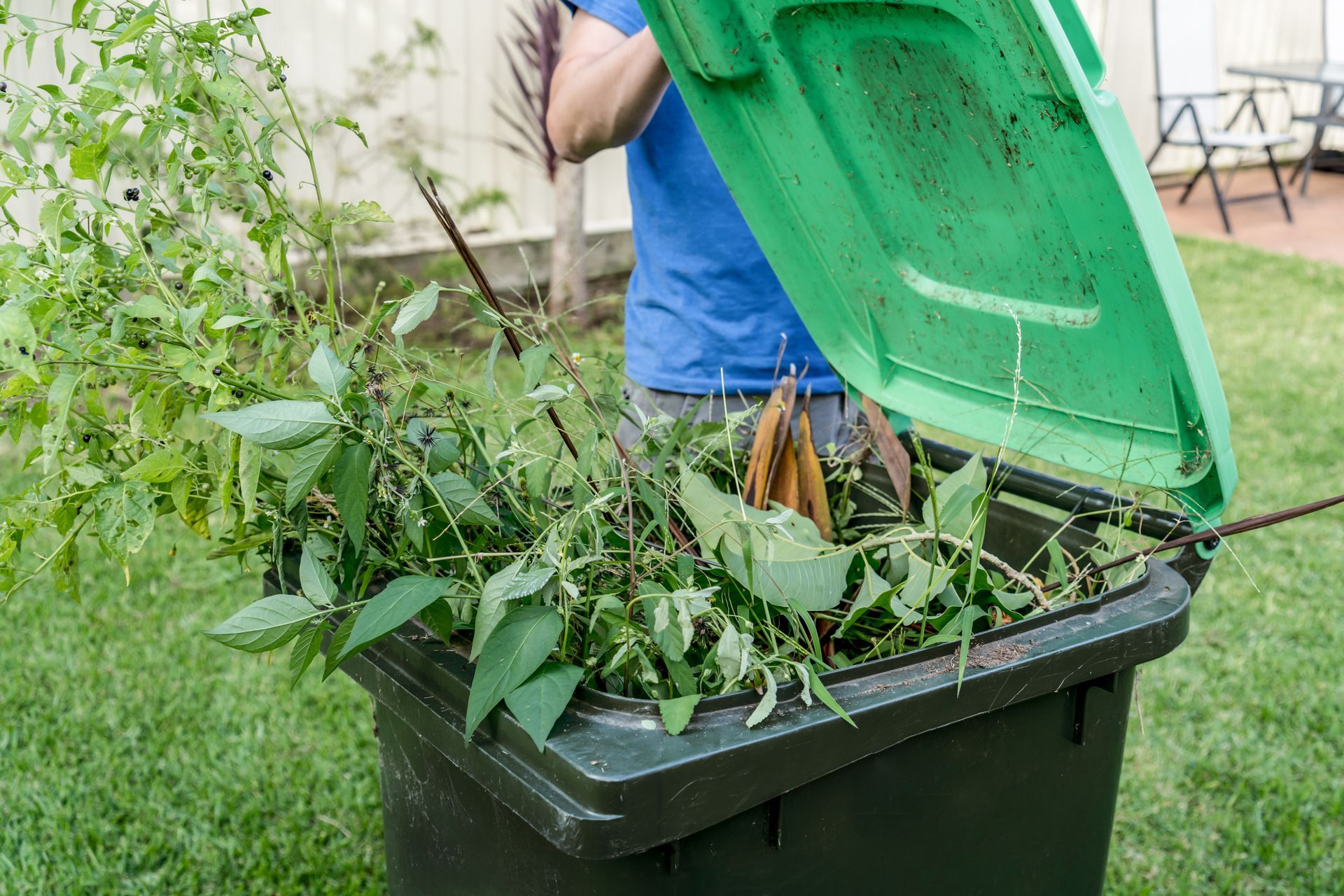 Person emptying green yard-waste bin into larger trash bin outdoors
