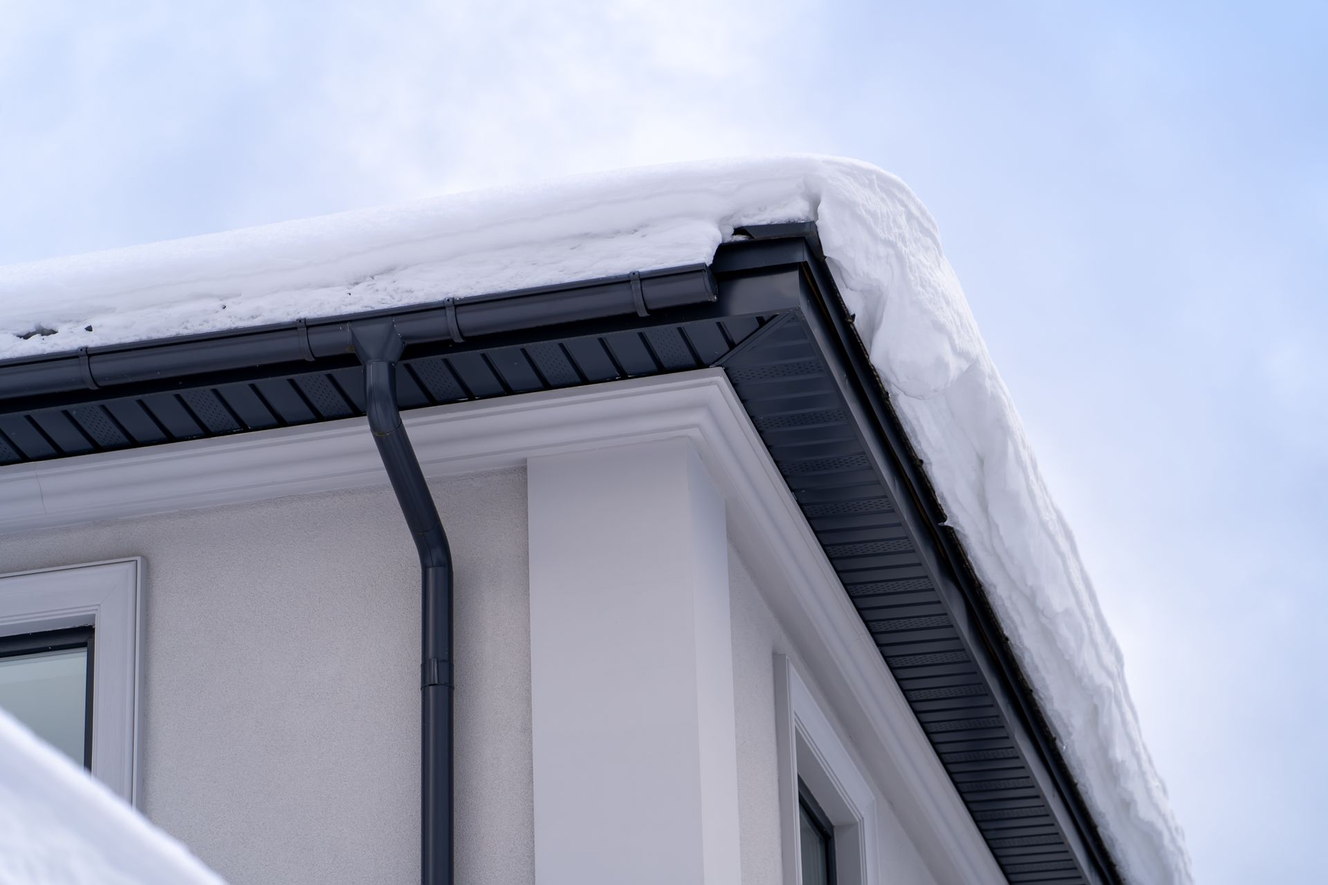 Snow-covered roof eaves and gutter on a house against a pale blue sky