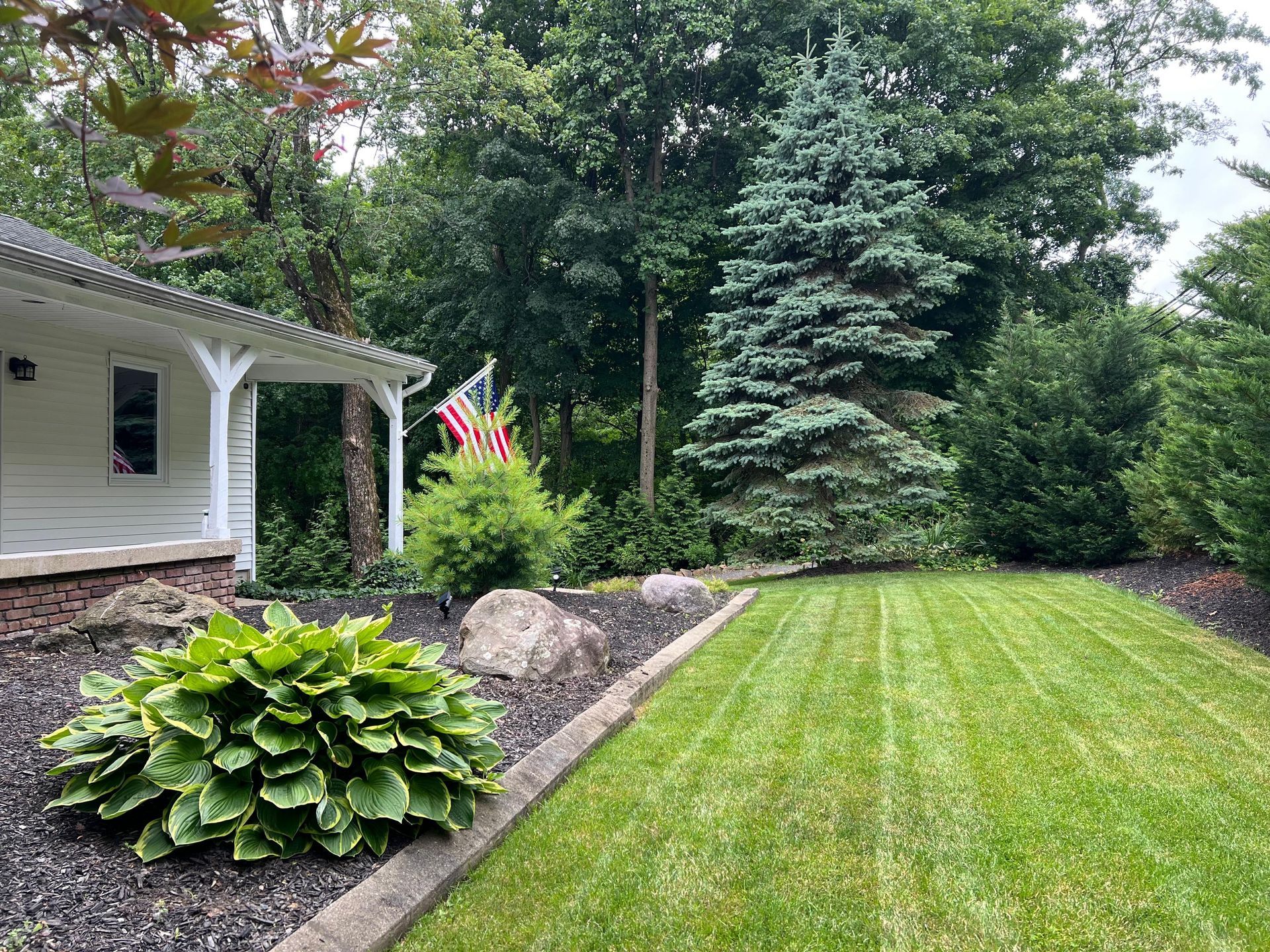 Well-kept front yard with green lawn, hostas, flower bed, and a porch beside tall trees