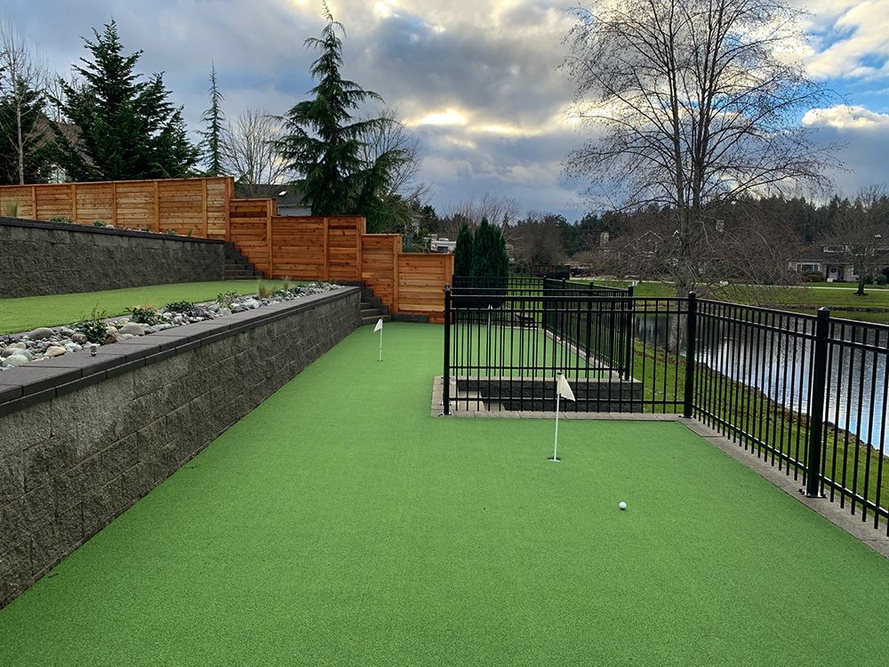 Bright green artificial turf walkway beside a fenced waterway and retaining wall under a cloudy sky