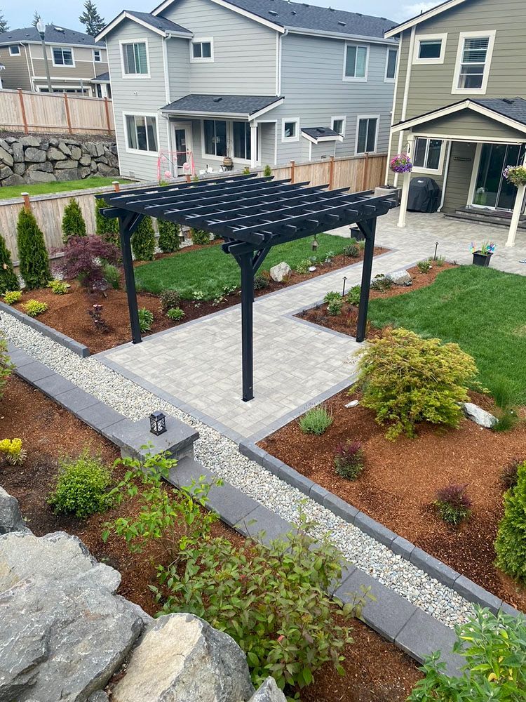Paved patio with black pergola, surrounded by landscaped garden and green lawn.