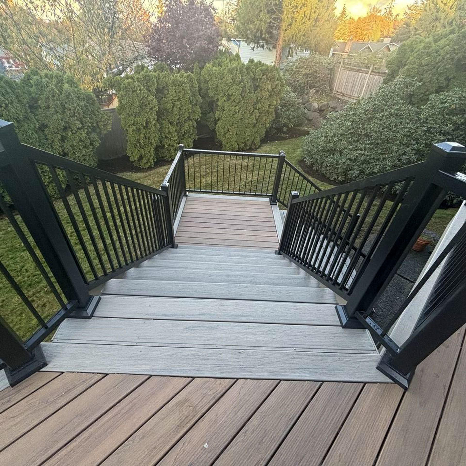 Wooden deck stairs with black railings leading down to a lush green backyard at sunset