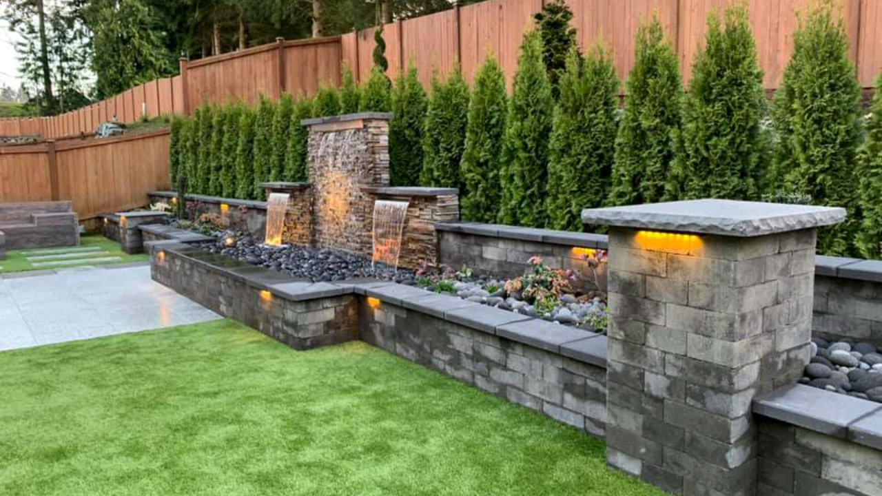 Backyard water feature with stone wall, waterfalls, and lush green lawn.